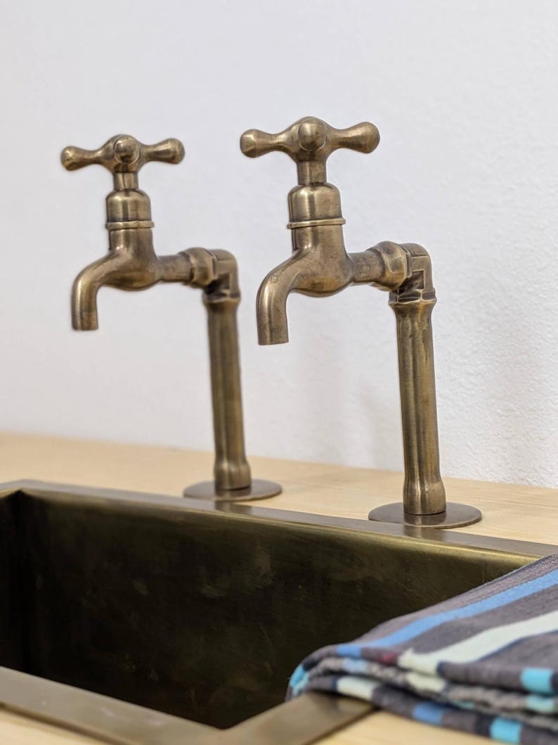 Puede incluir: Dos grifos de lat&oacute;n de estilo antiguo con mandos en cruz montados sobre una superficie de madera clara. Los grifos presentan un acabado met&aacute;lico desgastado y est&aacute;n situados detr&aacute;s de un lavabo rectangular de lat&oacute;n con un pa&ntilde;o a rayas.