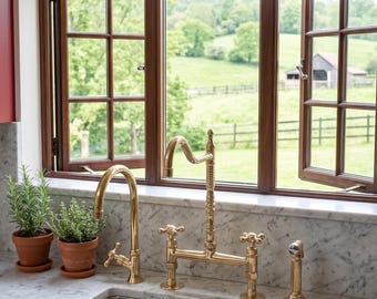 Puede incluir: Un fregadero de cocina con un grifo dorado y una ventana con vistas a un paisaje verde. El fregadero está situado en una encimera de mármol, con hierbas en macetas y un plato de fresas cerca. Una lámpara de estilo vintage está montada encima de la ventana.