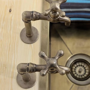 Puede incluir: Dos grifos de metal en tono lat&oacute;n antiguo con manijas en forma de cruz montados sobre una superficie de madera clara. Los grifos presentan una construcci&oacute;n similar a una tuber&iacute;a con placas base circulares sobre un lavabo met&aacute;lico.