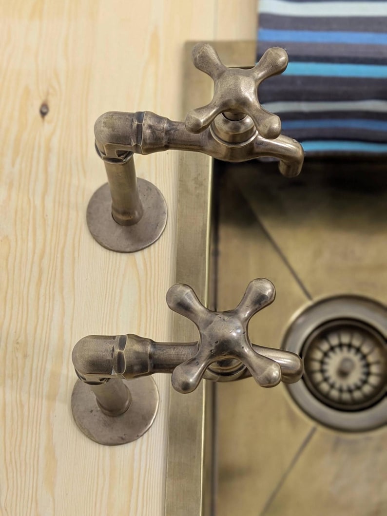 Puede incluir: Dos grifos de metal en tono lat&oacute;n antiguo con manijas en forma de cruz montados sobre una superficie de madera clara. Los grifos presentan una construcci&oacute;n similar a una tuber&iacute;a con placas base circulares sobre un lavabo met&aacute;lico.