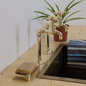 May include: Two brass faucets with cross handles are mounted on a light wood surface. A matching brass soap dish holds a bar of soap. A small potted plant and a striped towel are also visible.