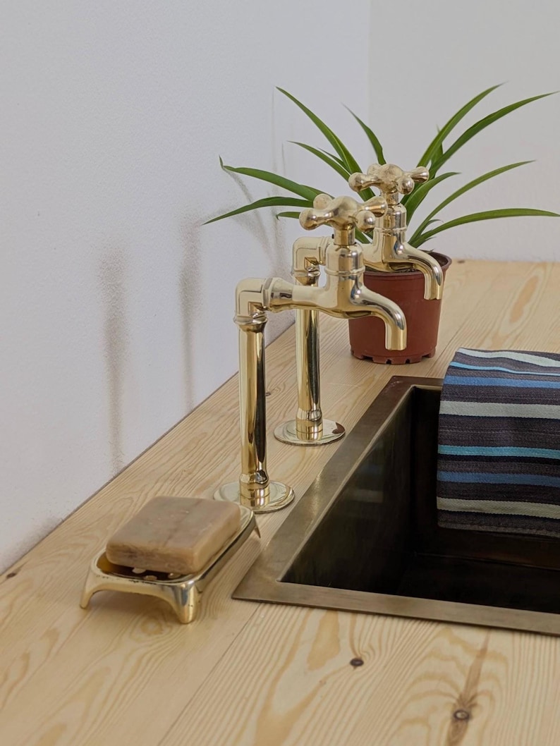 May include: Two brass faucets with cross handles are mounted on a light wood surface. A matching brass soap dish holds a bar of soap. A small potted plant and a striped towel are also visible.