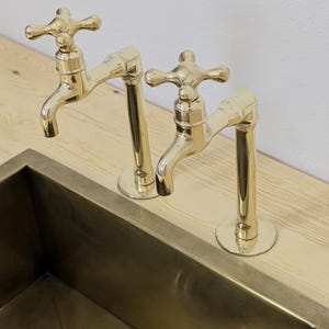 May include: Two brass faucets with cross handles are mounted on a light wood surface, positioned above a rectangular brass sink. The faucets have a classic design and a polished finish, reflecting the surrounding light.