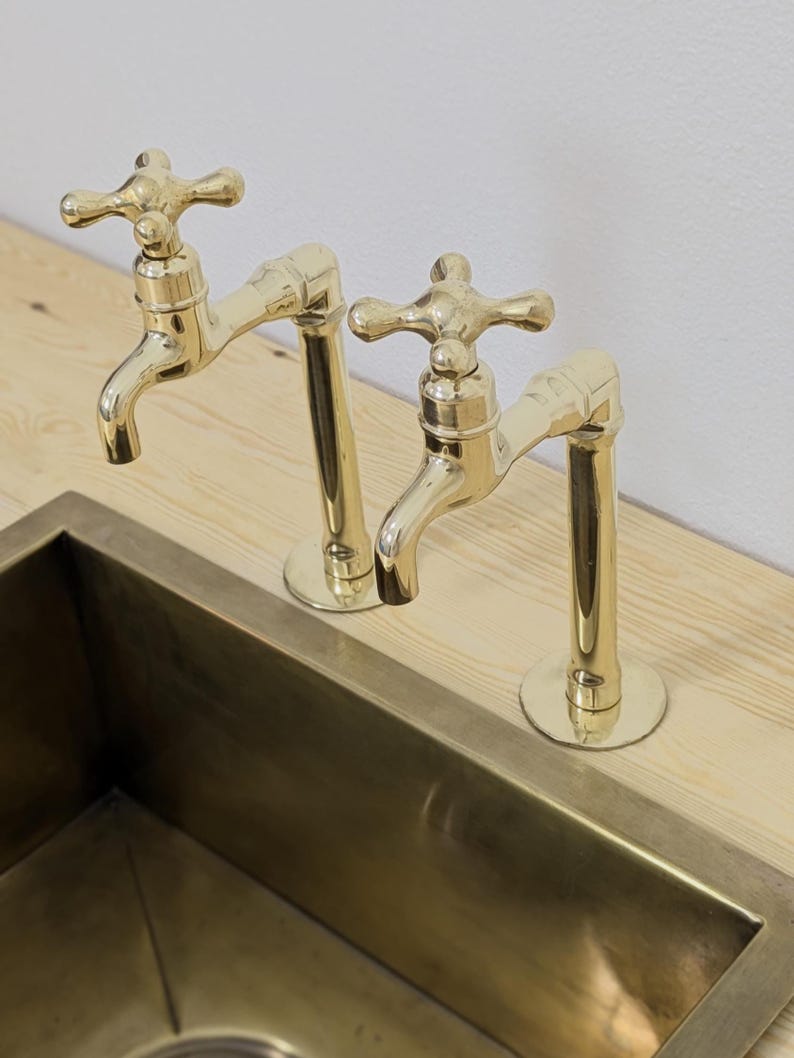 May include: Two brass faucets with cross handles are mounted on a light wood surface, positioned above a rectangular brass sink. The faucets have a classic design and a polished finish, reflecting the surrounding light.