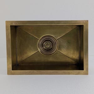 Op de afbeelding: Een rechthoekige messing spoelbak met een centrale afvoer. De spoelbak heeft een geborstelde afwerking en een decoratieve afvoerafdekking. De binnenkant van de spoelbak loopt schuin af naar de afvoer. De spoelbak staat tegen een witte achtergrond.