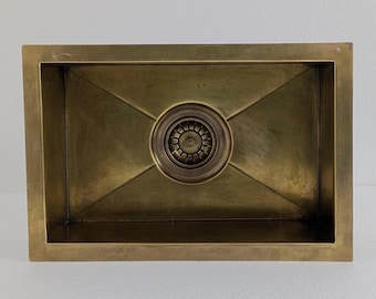 Puede incluir: Fregadero de cocina rectangular de latón con desagüe central. El fregadero tiene un acabado cepillado y una tapa de desagüe decorativa. El interior del fregadero se inclina hacia el desagüe. El diseño general es moderno y minimalista.