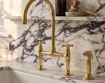 Puede incluir: Un grifo de cocina de oro pulido con caño de arco alto y dos mandos en cruz, acompañado de un rociador lateral a juego. El accesorio está montado sobre una encimera de mármol blanco con vetas moradas, sobre un fregadero de color blanco.