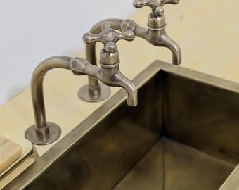 May include: A rectangular brass sink basin with two matching brass cross-handle faucets mounted on a light wood surface. The sink features a central drain and a metallic finish, accompanied by a blue and gray striped cloth resting on the edge.