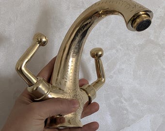 Puede incluir: Un grifo dorado con un caño curvo y dos manijas de palanca. El grifo tiene un diseño ornamentado con detalles grabados. El caño tiene un aireador negro. El grifo se sostiene en una mano.