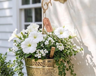 Puede incluir: Una maceta colgante de metal dorado con un patrón en relieve y un borde de greca, suspendida de un soporte de pared ornamentado a juego. La maceta contiene petunias blancas y follaje verde colgante, con una pequeña etiqueta de marca.