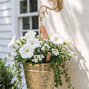 May include: A gold-colored hanging planter filled with white petunias and small white flowers. The planter is suspended from a decorative wall hook. The planter has an embossed design and trailing green vines.