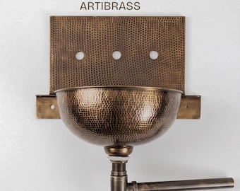 Puede incluir: Un lavabo de pared de latón martillado con una placa trasera rectangular con tres orificios circulares y un cuenco semiesférico. La unidad incluye un conjunto de tubería de drenaje de latón a juego que se extiende hacia la derecha. El texto ARTIBRASS es visible.