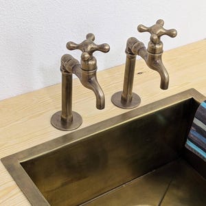 Puede incluir: Dos grifos de lat&oacute;n de estilo antiguo con manijas en forma de cruz montados sobre una superficie de madera clara junto a un fregadero rectangular de lat&oacute;n. Los grifos presentan un ca&ntilde;o curvo y un acabado met&aacute;lico a juego con el desag&uuml;e.