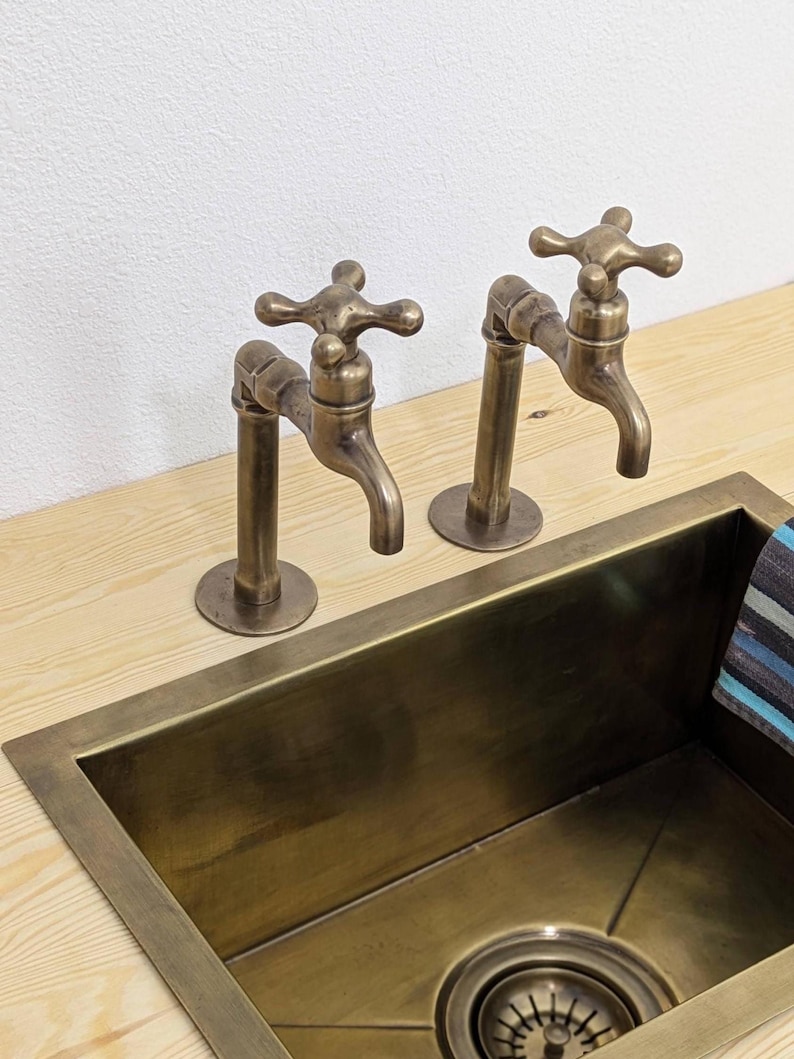 Puede incluir: Dos grifos de lat&oacute;n de estilo antiguo con manijas en forma de cruz montados sobre una superficie de madera clara junto a un fregadero rectangular de lat&oacute;n. Los grifos presentan un ca&ntilde;o curvo y un acabado met&aacute;lico a juego con el desag&uuml;e.