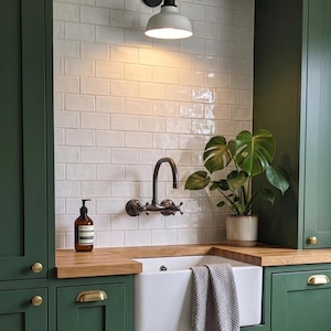 Può includere: Una cucina con mobili verde scuro, un lavello a vasca bianco e un piano di lavoro in legno. Un rubinetto in stile vintage e una pianta in vaso sono visibili. Un paraschizzi piastrellato bianco e un'applique in bianco e nero completano la scena.
