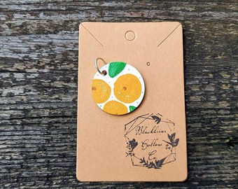 May include: A round wooden ornament with a white background and a painted design of orange slices and green leaves. The ornament has a silver loop for hanging and is labeled "Blackbird Hollow Co."