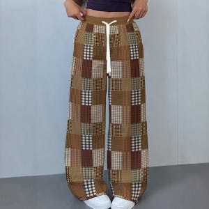 以下が含まれることがあります： 茶色、ベージュ、クリーム色のワイドレッグパッチワークパンツ。 パンツは、ドローストリングウエストと、さまざまなテクスチャとプリントの正方形の繰り返しパターンが特徴です。 モデルは白いスニーカーを履いています。