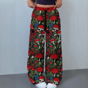 May include: Wide-leg trousers featuring a vibrant all-over print of red mushrooms and green foliage on a black background. The trousers have a solid red elastic waistband with a black drawstring cord and are paired with white trainers.