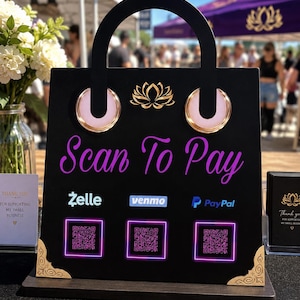 May include: A black, shopping bag-shaped payment sign with a handle, featuring the purple text Scan To Pay, a gold lotus icon, and three QR codes. It displays the Zelle, Venmo, and PayPal logos, with gold-colored decorative corner accents.
