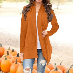 Könnte beinhalten: Ein orangefarbener Cardigan mit langen Ärmeln. Der offene Pullover hat eine warme, einfarbige Farbe. Das Model trägt blaue Jeans mit zerrissenen Patches. Kürbisse sind im Hintergrund.
