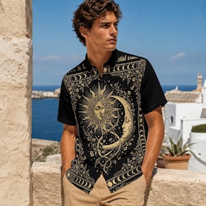 May include: A black short-sleeved button-down shirt featuring a gold-toned graphic print of a sun and crescent moon surrounded by celestial motifs and floral patterns. The design includes the text "Celestial Dance" on the upper chest area.