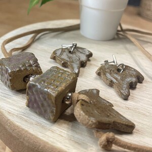May include: A brown and beige ceramic necklace with a variety of charms, including a square charm, a leaf charm, and a bird charm. The necklace is strung on a brown cord.