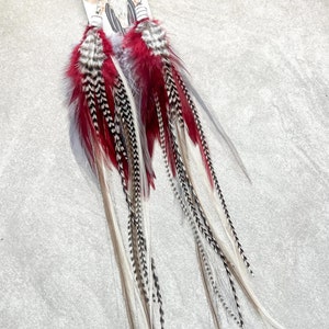 May include: A pair of long feather earrings with red, black, white, and gray feathers. The earrings are attached to a white card with the text "Fringed in Faith" and a graphic of a skull and feathers.