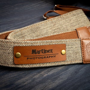 May include: A brown and beige camera strap with a brown leather patch that says "Martinez Photography" in black lettering. The strap has a herringbone pattern and two brass rivets.