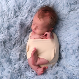 May include: A newborn baby is swaddled in a cream-colored fabric and sleeping on a light blue fluffy blanket.