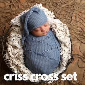 May include: A newborn baby is swaddled in a blue criss-cross wrap and a matching blue knitted hat. The baby is sleeping in a white crocheted basket.