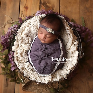 May include: A newborn baby girl is sleeping peacefully in a purple swaddle with a white lace trim. She is wearing a white headband with a small flower accent. The baby is lying in a metal basket lined with a white crocheted blanket. The basket is surrounded by purple flowers.