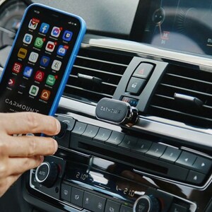 May include: A black magnetic phone mount attached to a car's air vent. A blue phone case with a smartphone inside is held in place by the mount. The phone screen displays various app icons.