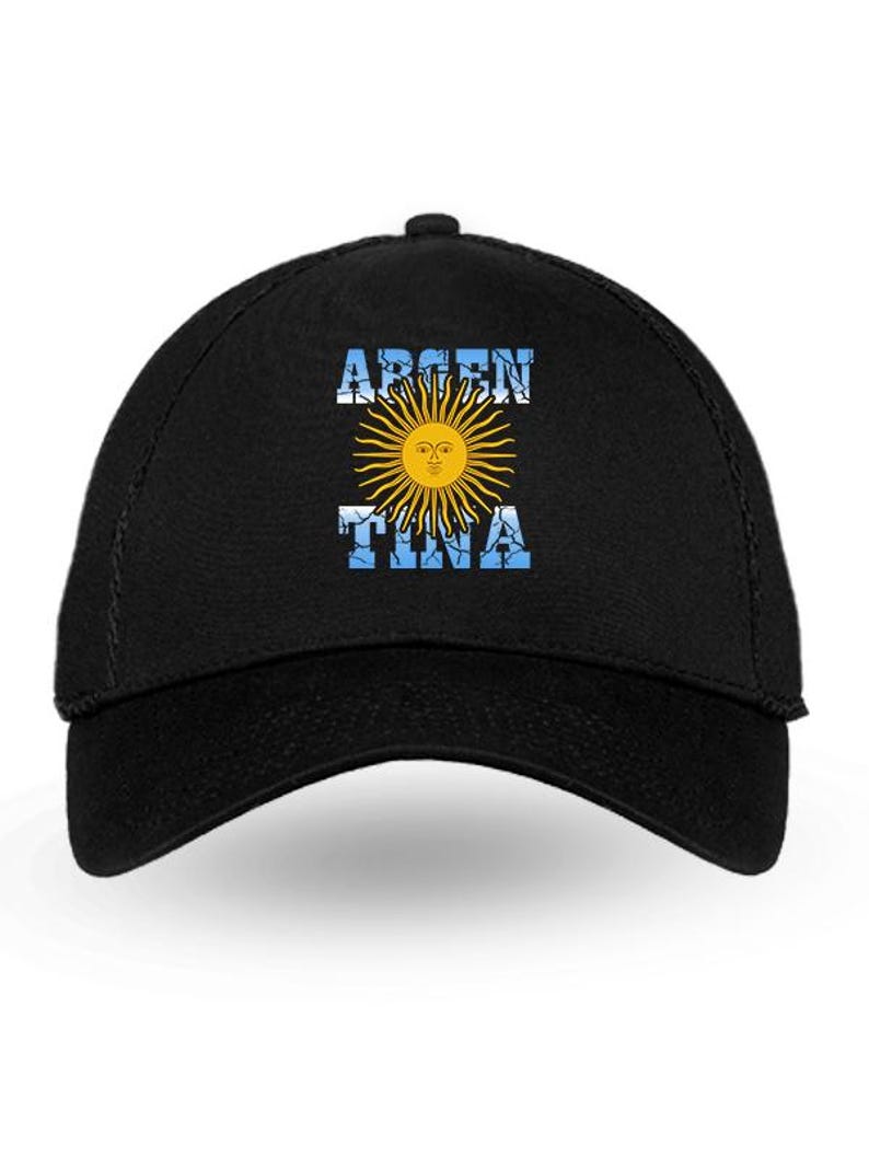 May include: Black baseball cap featuring the word "ARGENTINA" in white, with a yellow sun design in the center. The sun has a face and is surrounded by rays. The background of the text is blue.
