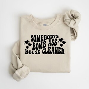 May include: A beige sweatshirt with the text "Somebody's Bomb Ass House Cleaner" in black, retro-style font. The text is surrounded by small black hearts.