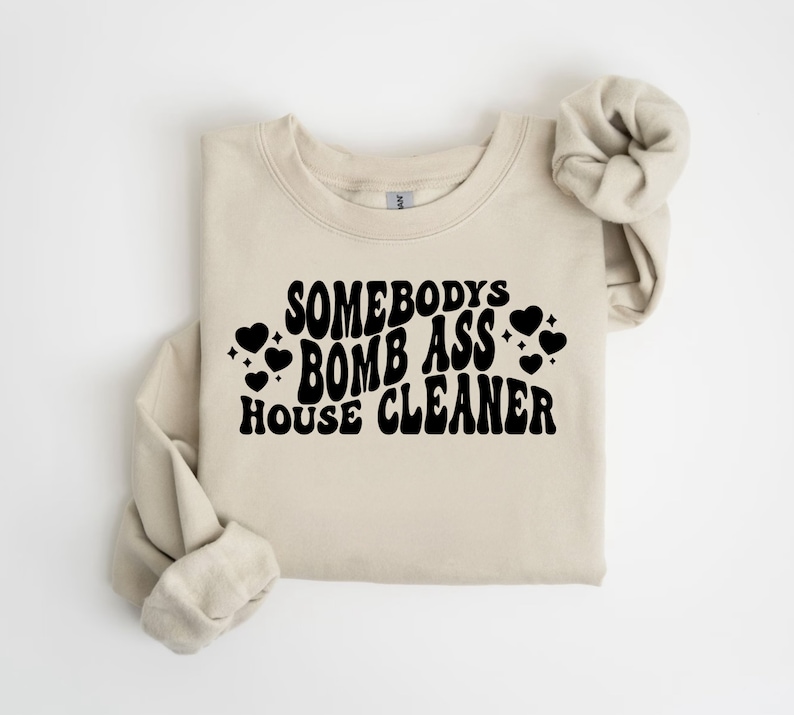 May include: A beige sweatshirt with the text "Somebody's Bomb Ass House Cleaner" in black, retro-style font. The text is surrounded by small black hearts.