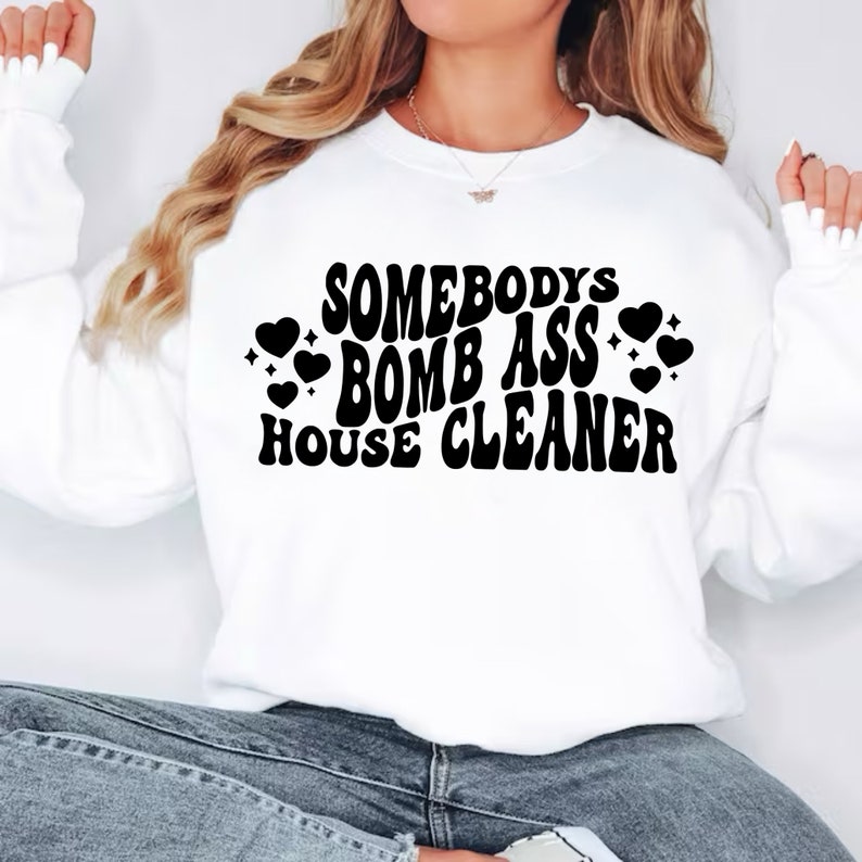 May include: White sweatshirt with black text that reads "Somebody's Bomb Ass House Cleaner" with hearts around the text.