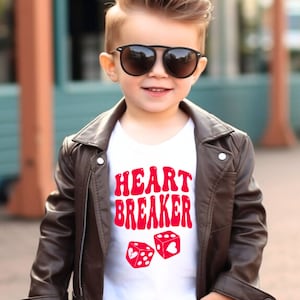 Op de afbeelding: Een kind draagt een wit t-shirt met de rode tekst "HEART BREAKER" en twee rode dobbelstenen. Het kind draagt ook een bruine leren jas en een zwarte zonnebril.