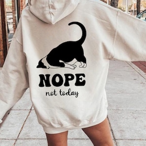以下が含まれることがあります： クリーム色のパーカー。黒と白の猫のイラストが描かれています。猫のグラフィックの下には「NOPE not today」というテキストがプリントされています。パーカーにはドローストリングフードとカンガルーポケットが付いています。