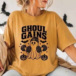 May include: A mustard yellow short-sleeve t-shirt featuring a black graphic print of a ghost wearing a beanie labelled BOO, lifting two dumbbells. The design includes the text GHOUL GAINS, two jack-o'-lanterns, bats, and small star accents.