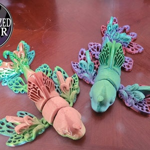 May include: Two 3D printed fish with intricate butterfly wings. One fish is green with purple wings, the other is orange with green wings.