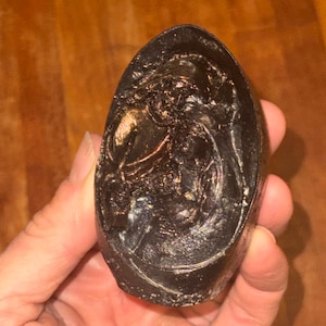 May include: A dark brown oval-shaped soap bar with a raised design of a person holding a staff. The soap has a shiny, almost metallic finish.