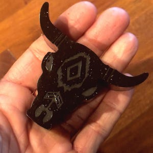 May include: A black bull head-shaped pin with a glossy finish. The pin features a diamond and arrow design on the head. The horns curve upwards. The pin is held in a hand.