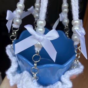 May include: A blue ceramic bowl with a white crocheted trim and hanging white cords. The cords are adorned with white pearl beads and small white satin bows. Silver-colored chain links connect the cords to the bowl.