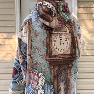 May include: A woven tapestry hoodie featuring a large grandfather clock graphic with a beige face and black numerals. The fabric displays a light blue background with pink rose patterns, a dark brown clock frame, and fringe detailing on the sleeves.