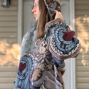 Puede incluir: Una chaqueta con capucha de tapiz que presenta un diseño tejido de osos de peluche, patrones florales y motivos de corazones en tonos azul, marrón y crema. La prenda tiene una construcción de tela texturizada con flecos en los puños y la capucha.