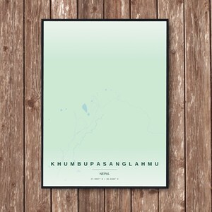 May include: A framed print featuring a map of Khumbu Pasang Lhamu, Nepal, in shades of green and white. The text "KHUMBU PASANGLAHMU" is at the bottom, with "NEPAL" below it, and coordinates. The print is displayed against a wooden background.