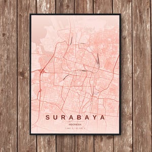 Op de afbeelding: Een ingelijste kaartafdruk van Surabaya, Indonesië, in koraaltinten. De stadskaart toont gedetailleerde straatindelingen en is gelabeld met de stadsnaam en het land. De print wordt weergegeven tegen een houten achtergrond.