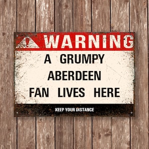 May include: A warning sign with a red triangle and the text "WARNING" in red letters. The sign reads "A GRUMPY ABERDEEN FAN LIVES HERE" in black letters. The sign also has the text "KEEP YOUR DISTANCE" in smaller black letters.