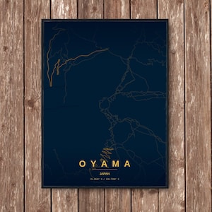 May include: A framed print featuring a map of Oyama, Japan, in gold lines against a dark blue background. The city name is displayed in gold letters, with the country and coordinates below. The print is displayed on a wooden surface.