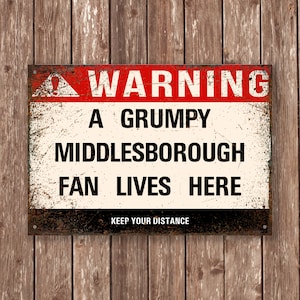 May include: A rectangular metal sign with a distressed finish, featuring the text "WARNING A GRUMPY MIDDLESBOROUGH FAN LIVES HERE" in bold black letters. The top border is red, and the bottom reads "KEEP YOUR DISTANCE."